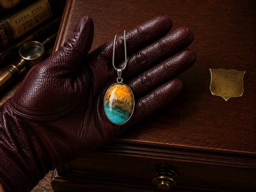Leather-gloved hand holding The Torchbearer Twelve Specimen Four, a one-of-a-kind wearable copper pendant necklace with bronze copper, turquoise verdigris and weathered landscape formations.